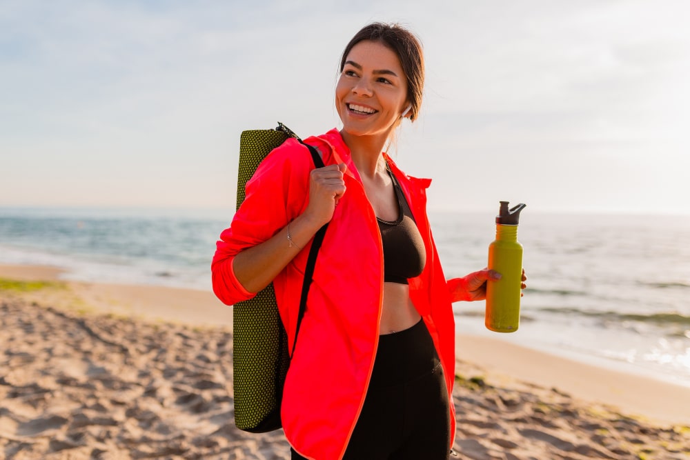 young-attractive-smiling-woman-doing-sports-in-morning-sunrise-on-sea-beach-holding-yoga-mat-and-bottle-of-water-healthy-lifestyle-listening-to-music-on-earphones-wearing-pink-windbreaker-jacket-min.jpg